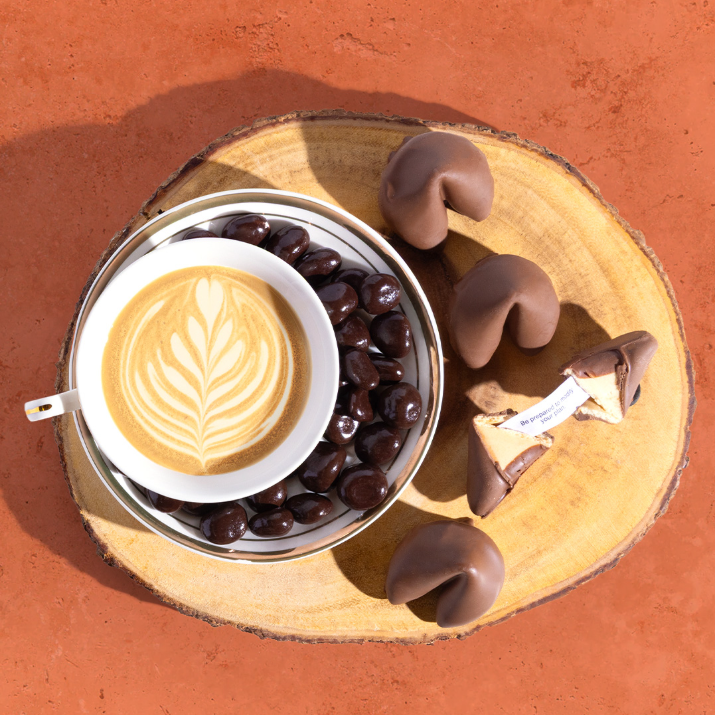 Cup of coffee with latte art, dark chocolate covered cherries, and milk chocolate covered fortune cookies on a wooden board.